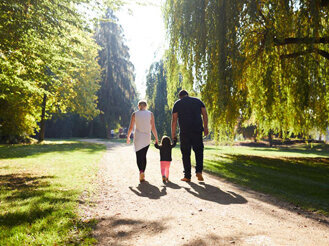 Auf dem Bild sieht man eine kleine Familie von hinten, die gerade einen langen, sonnengefluteten Weg durch einen Park nimmt.